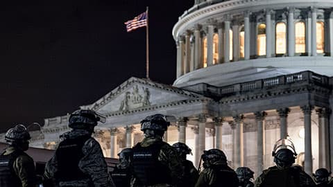 The image is from Elesa Zehndorfer's book Post-Truth Politics: A Brave New World?. There are eight people dress in army outfits outside a building with an american flag on it.